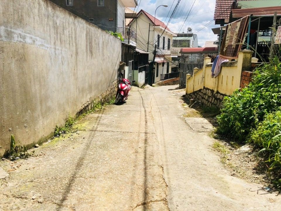 Con hẻm bê tông giữa một bức tường rào và dãy nhà, một xe máy đỏ dựng sát tường