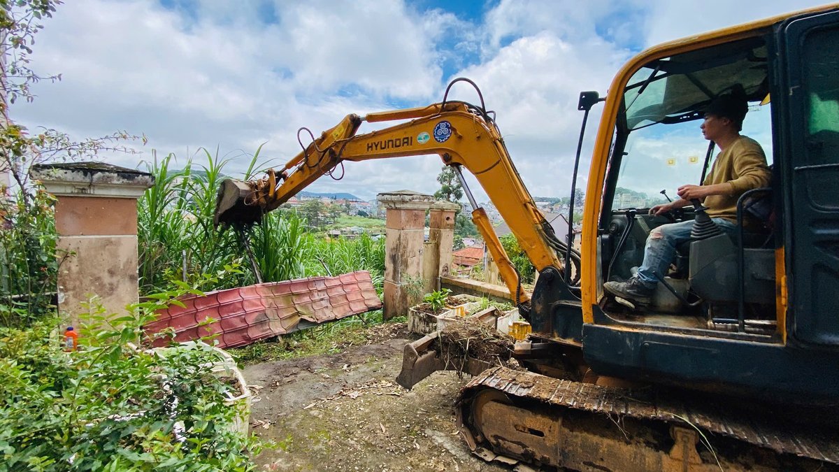 Người lái máy xúc dọn một lô đất có trụ cổng cũ, tấm tôn đổ và cỏ mọc cao