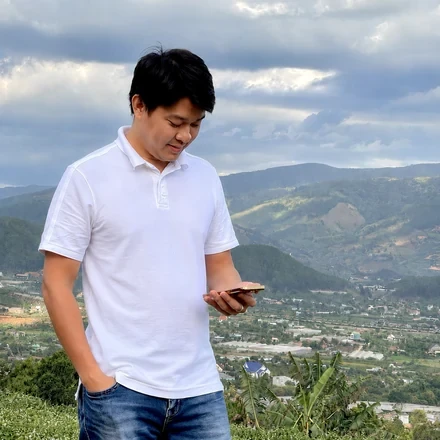 Đoàn Quốc Duyệt mặc áo polo trắng, cầm điện thoại, đứng trên đồi, phía sau là thung lũng có nhà cửa và dãy núi